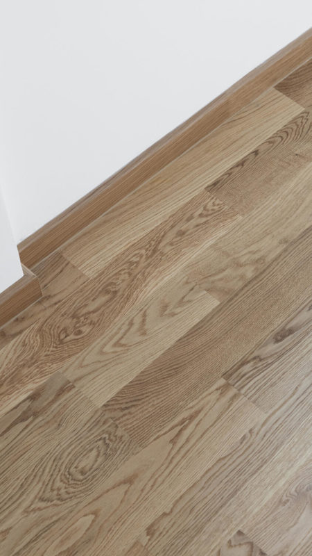 Паркетная доска Tarkett Oak Essence, крупный план укладки плашек натурального дуба под углом у светлой стены с плинтусом.