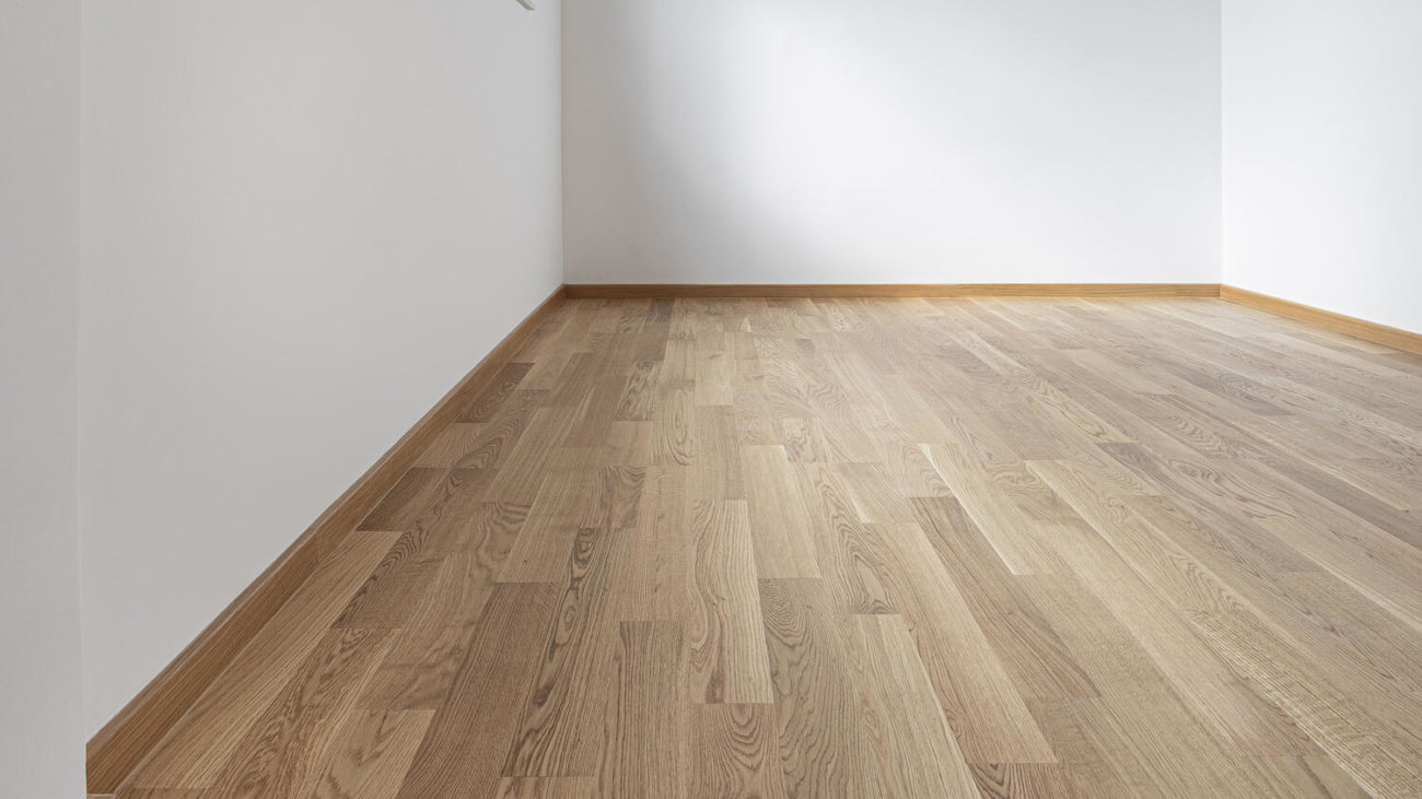 Двухполосная паркетная доска Tarkett Oak Essence в интерьере пустой светлой комнаты с белыми стенами и деревянным плинтусом.
