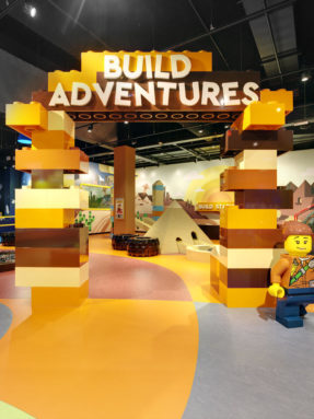 Оранжевый и коричневый линолеум Tarkett в интерьере зоны Build Adventures с большой аркой из блоков конструктора и фигуркой человечка.