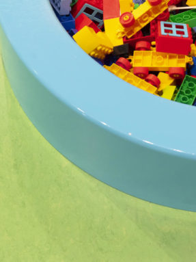 Текстура салатового линолеума Tarkett в детской игровой зоне LEGO рядом с круглым голубым бассейном, наполненным разноцветными кубиками.