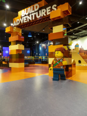 Серый и оранжевый линолеум Tarkett в интерьере зоны Build Adventures с большой аркой из кубиков LEGO и фигурой человечка на переднем плане.