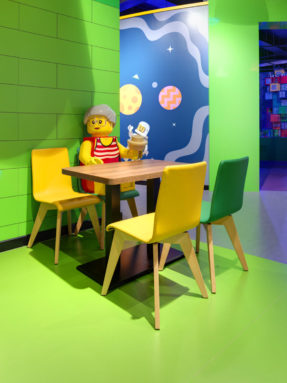 Салатовый линолеум Tarkett в интерьере детского кафе LEGO с фигурой человечка за обеденным столом и яркими цветными стульями.