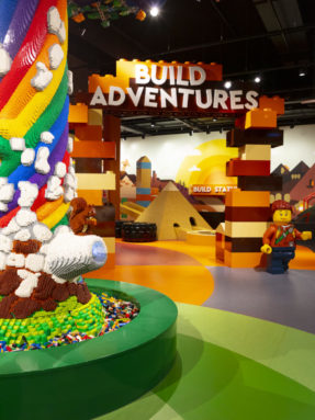 Разноцветный линолеум Tarkett в интерьере детской зоны Build Adventures с ярким радужным деревом из кубиков и фигуркой человечка.