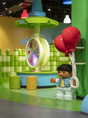 Линолеум Tarkett серого и зеленого цвета в интерьере детской зоны LEGO Duplo с интерактивной каруселью и большой фигурой мальчика.