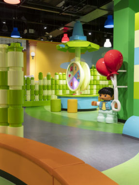 Зеленый и серый линолеум Tarkett в интерьере детской зоны LEGO Duplo с крупной фигуркой мальчика, воздушными шарами и игровыми элементами.