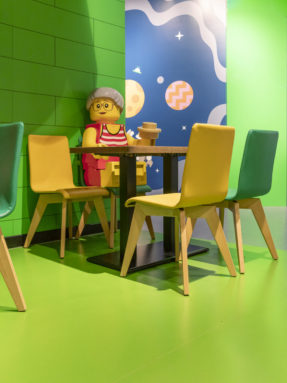 Зеленый линолеум Tarkett в интерьере игровой зоны LEGO с обеденным столом, желтыми стульями и большой фигурой человечка.