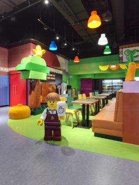 Линолеум Tarkett серого и салатового цвета в интерьере детского кафе LEGO с фигурой повара и деревянными обеденными столами.