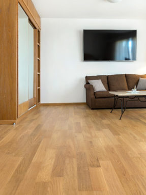 Паркетная доска Real Wood GR Kydon Hotel 08 натурального оттенка дуба в интерьере гостиной с коричневым диваном и телевизором на стене